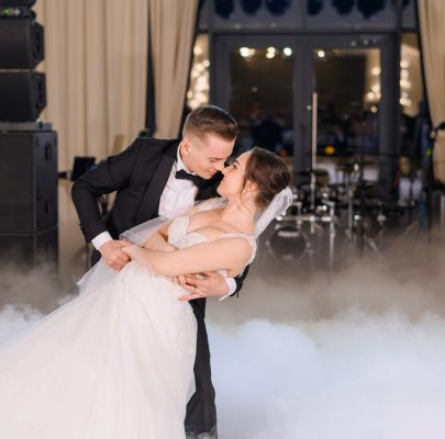 Side view of romantic moment of married couple dancing in smoke and groom titling elegant bride in long dress, mman and woman celebrating wedding party in restaurant
