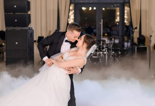 Handsome Groom Holding Bride While Dancing Wedding Dance Side view of romantic moment of married couple dancing in smoke and groom titling elegant bride in long dress, mman and woman celebrating wedding party in restaurant